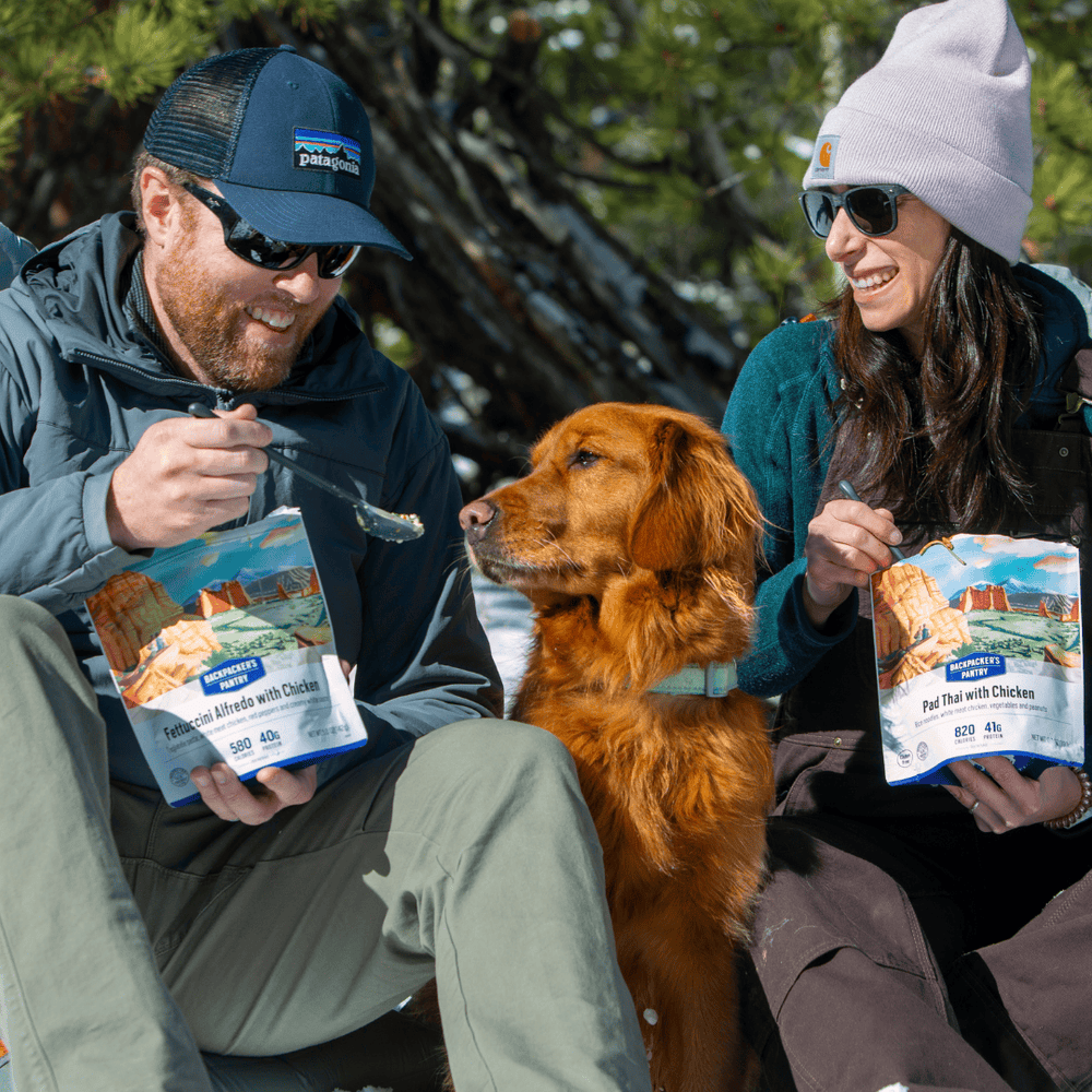 Two people eating camping food with a dog
