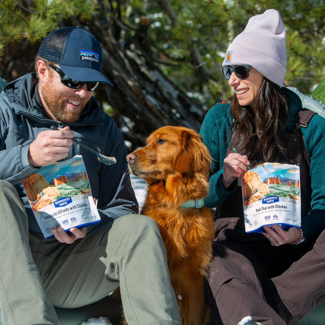 Two people sitting outdoors with a dog, holding freeze dried pouches