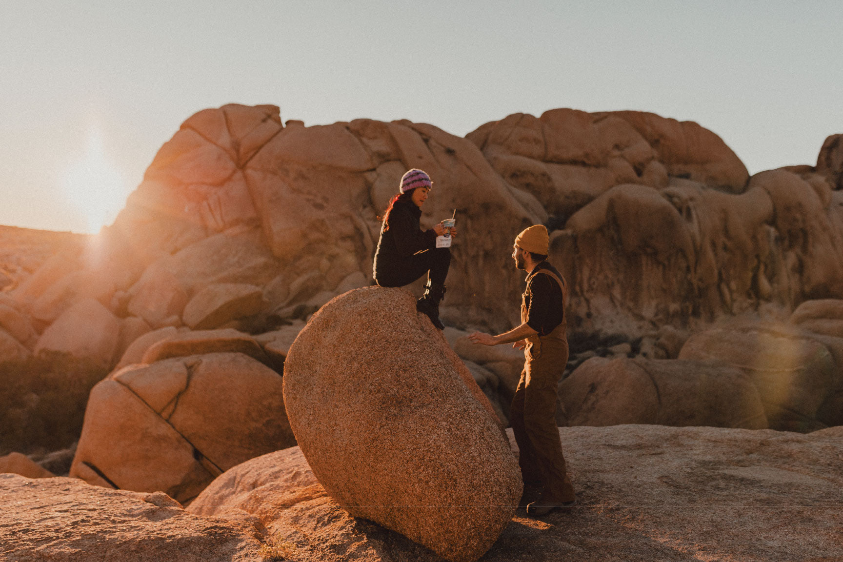 Two people sitting on large rocks in a desert landscape with a sunset.